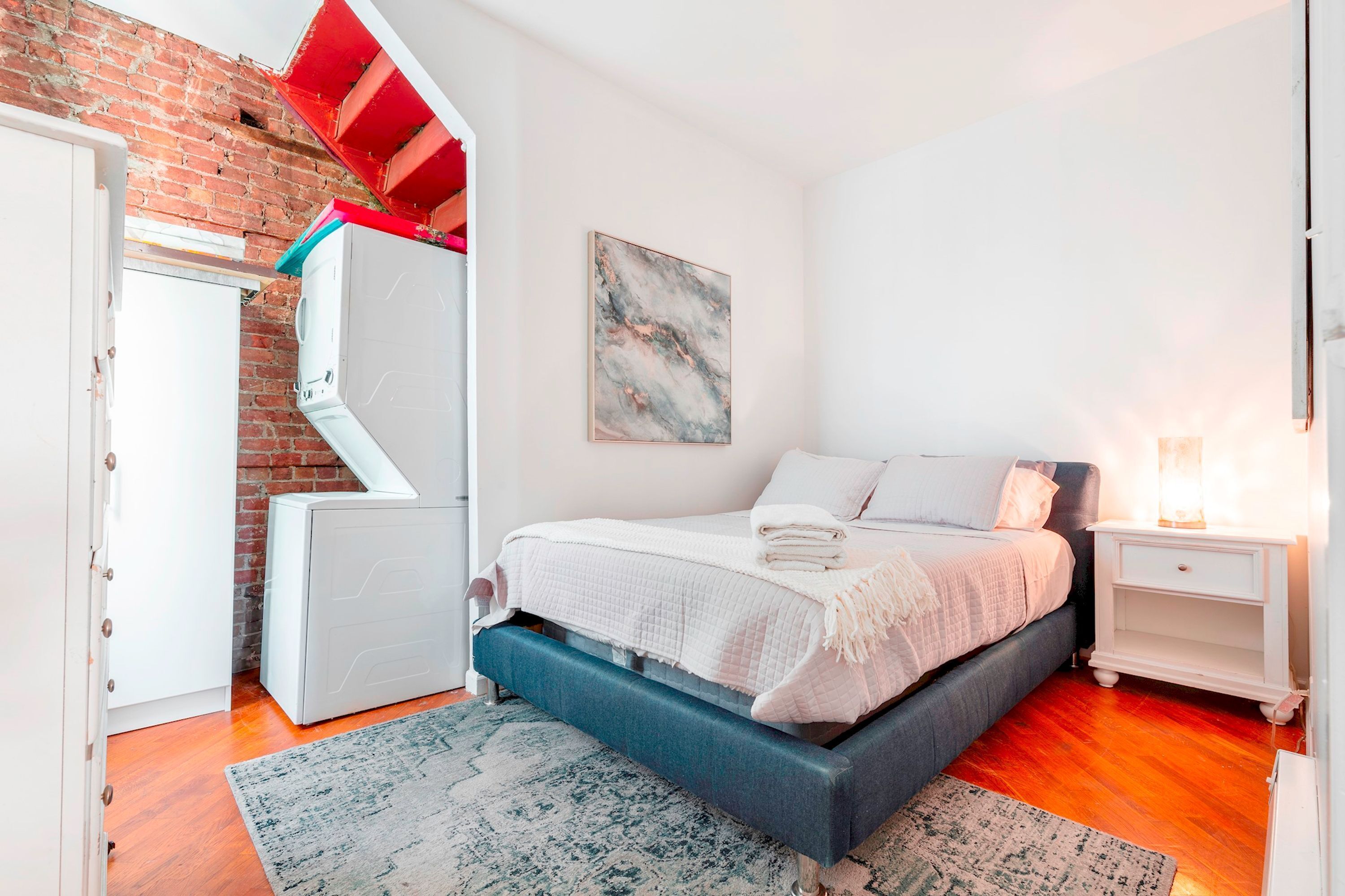 Een slaapkamer met een bed, nachtkastje, een witte dressoir, een rode en witte kast en een bakstenen muur.