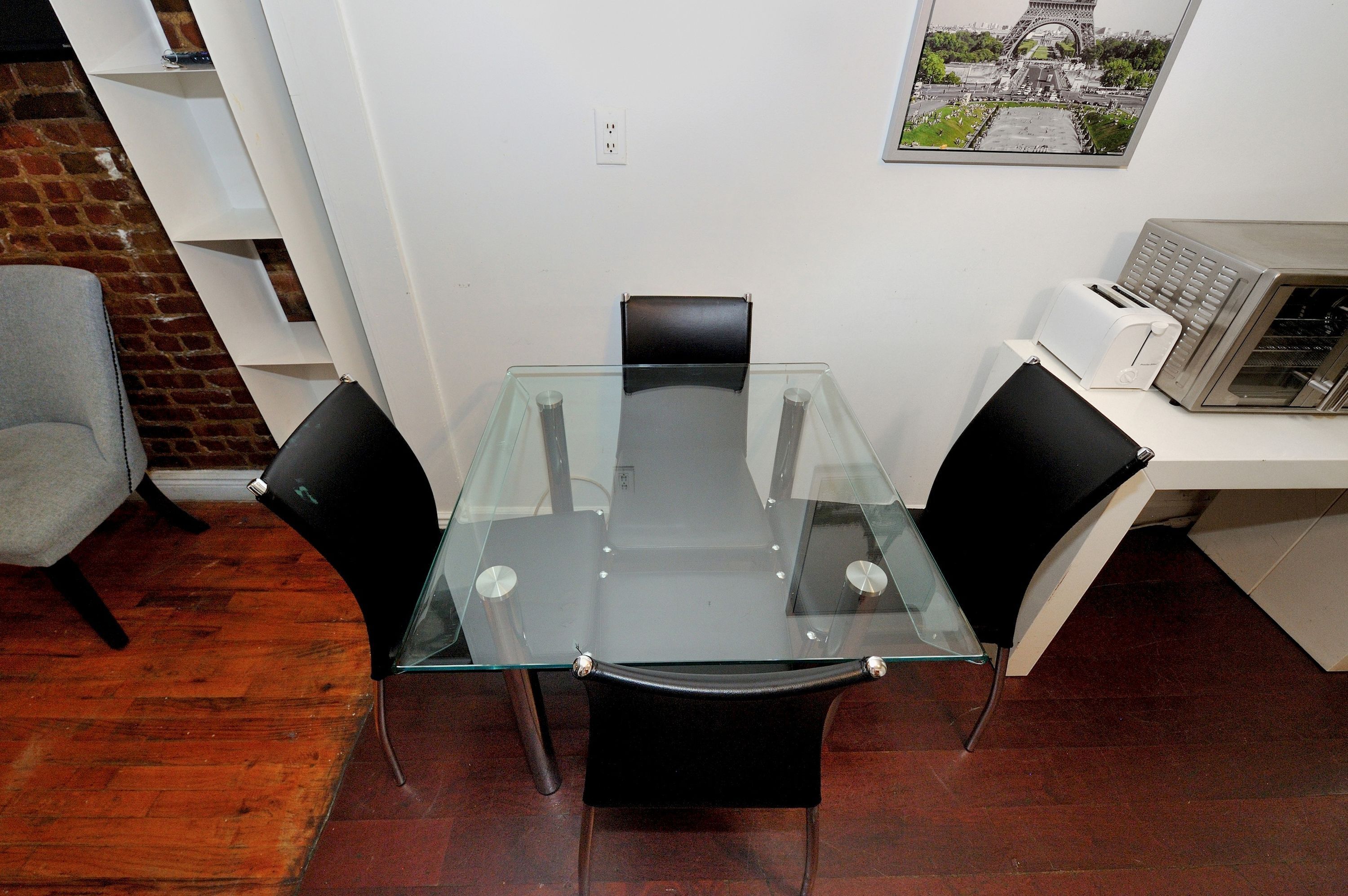 Une table à manger avec un plateau en verre et quatre chaises, un mur en briques, un mur blanc orné d’un tableau, et un four à micro-ondes posé sur un comptoir blanc.