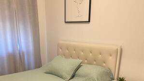 Een netjes opgemaakt bed met een lichtgroene spreidek, twee kussens, een witte nachtkastje met een plantje en een lijst met een kunstwerk aan de muur.