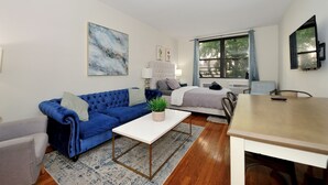 Ein modernes Wohnzimmer mit einem blauen Sofa, einem weißen Couchtisch und einem grauen Sessel. Ein Fenster bietet Blick auf Bäume, und ein Bett hat ein graues Kopfteil.