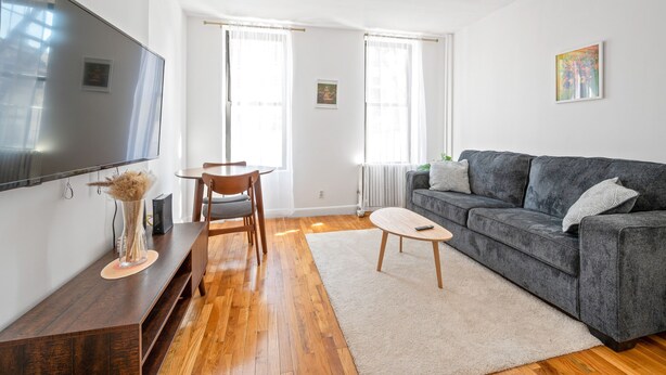 Una sala de estar moderna con un sofá gris, piso de madera, un televisor de pantalla plana, una mesa de comedor con sillas y ventanas amplias con cortinas ligeras.