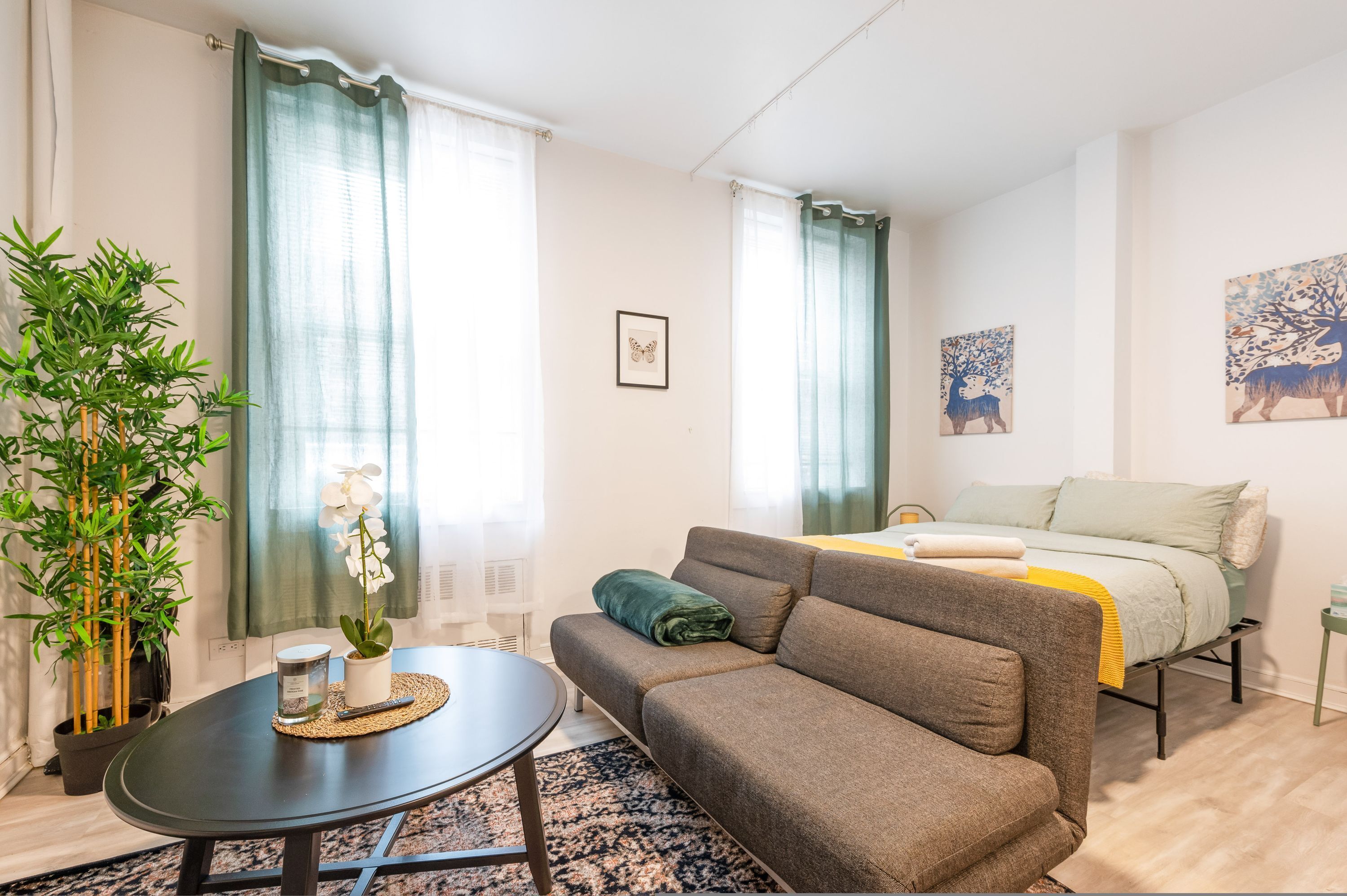 Ein modernes Wohnzimmer mit einer Couch, einem kleinen Tisch, einem Bett mit einer grünen und gelben Tagesdecke sowie einem Bild von Hirschen an der Wand.
