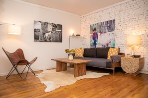 A living room with a wooden floor, a sofa with patterned cushions, a coffee table, a leather chair, and a wall with a textured design and two artworks. - Sunny Fully Furnished Studio in Chelsea (New York)