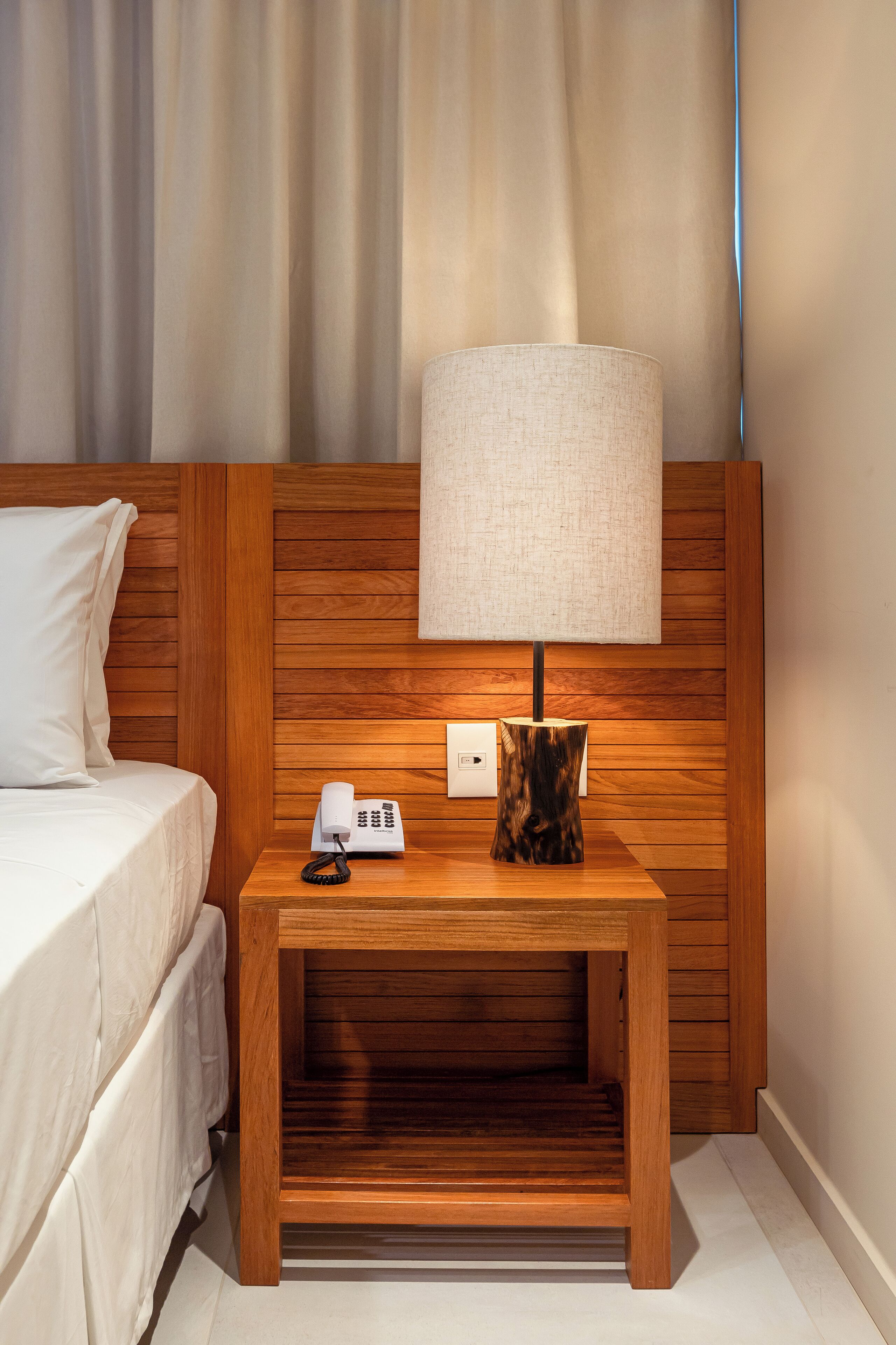 A wooden bedside table with a lamp, a telephone, and a power outlet.