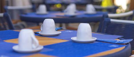 A row of tables with white cups and saucers, blue tablecloths, and metal chairs.