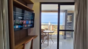 Una habitación con televisión mostrando varios canales, un balcón con mesa y sillas, y vistas al mar y edificios.