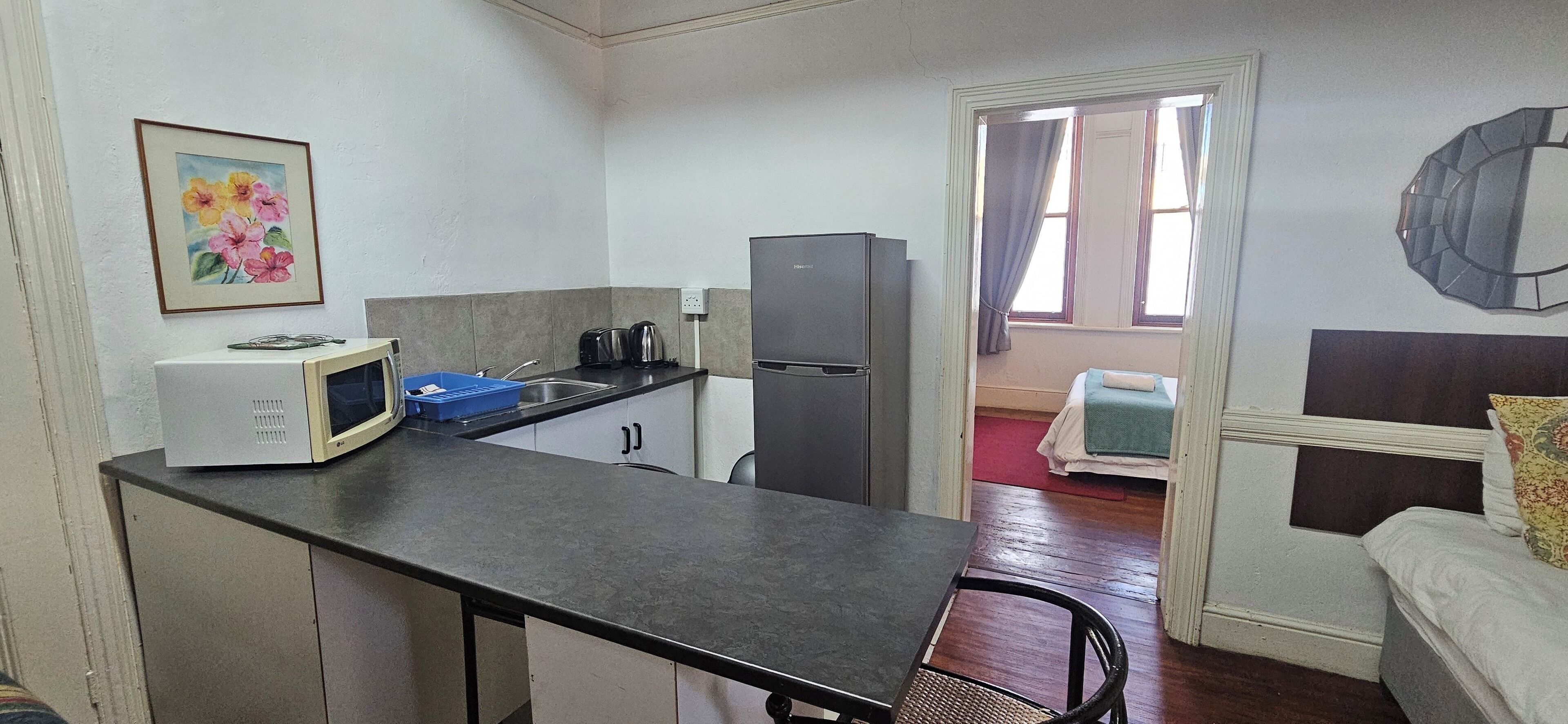 A compact kitchen with a microwave, toaster, and refrigerator. There is a small dining area with a chair and a table. A painting hangs on the wall, and a window with curtains is visible.