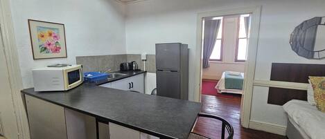A compact kitchen with a microwave, toaster, and refrigerator. There is a small dining area with a chair and a table. A painting hangs on the wall, and a window with curtains is visible.