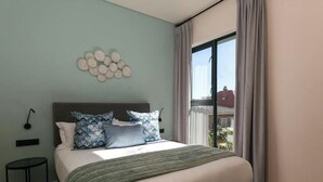 Ein Schlafzimmer mit einem Bett, einer Wandleuchte, einem kleinen Tisch und einem fenster mit Vorhängen.