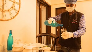 Una persona con uniforme de chef vertiendo un líquido en un vaso. Hay un reloj en la pared, una puerta y varios utensilios de cocina sobre la mesa.