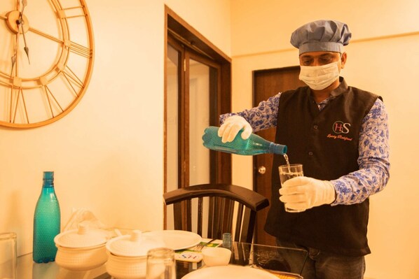 Une personne vêtue d’un uniforme de chef verse un liquide dans un verre. On peut voir une horloge au mur, une porte et divers ustensiles de cuisine sur la table.