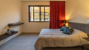 Quarto com cama, janela com cortinas, mesa de cabeceira com abajur e prateleira com toalhas.