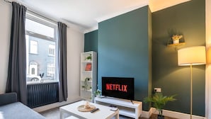 Una sala de estar con un sofá, una mesa de centro, un televisor sobre un mueble, una lámpara de pie y una ventana con cortinas.