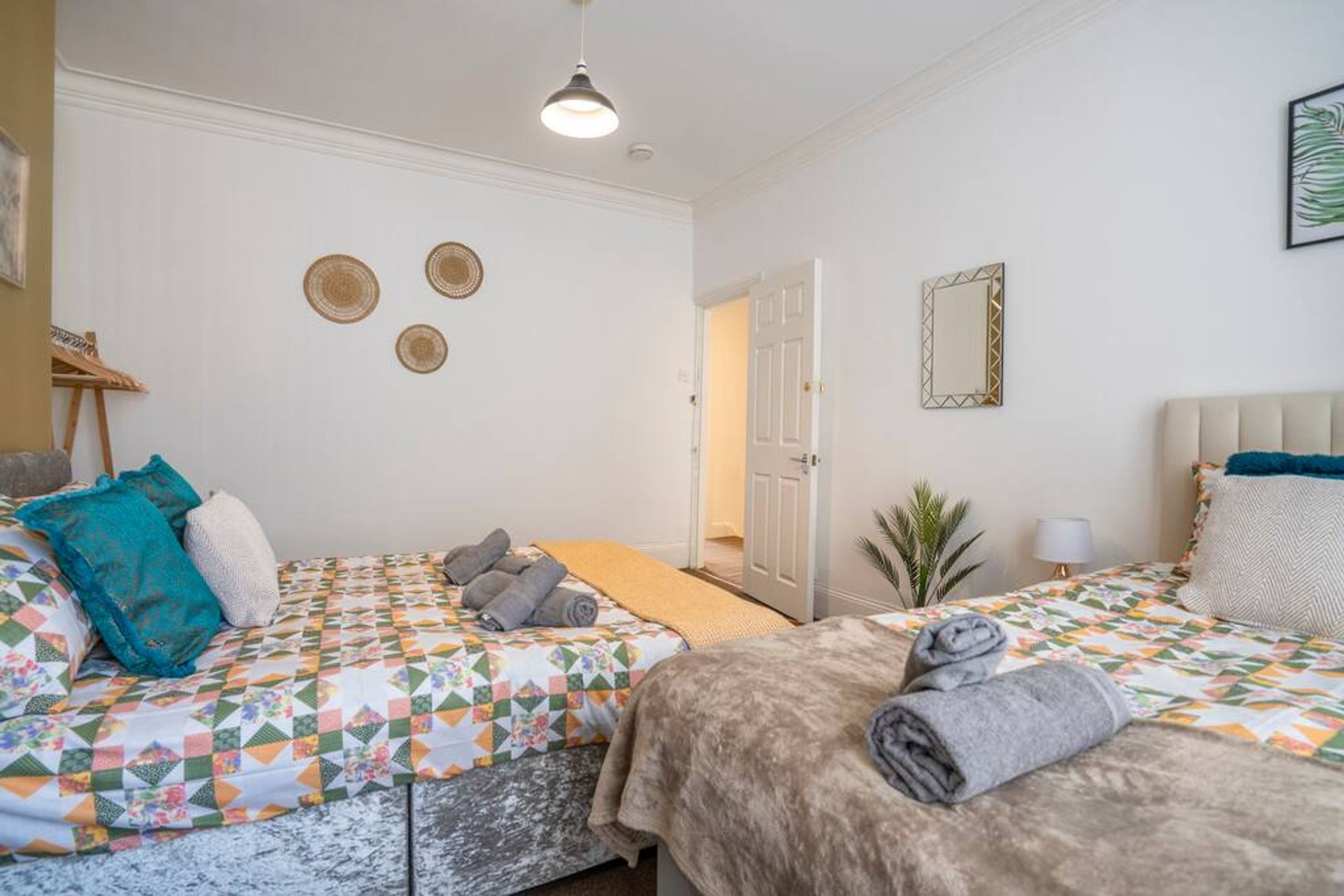 Ein Hotelzimmer mit zwei Betten, jeweils mit einem gemusterten Überwurf, einem kleinen Tisch, einem Stuhl, einer Lampe und einer Tür zu einem weiteren Raum.