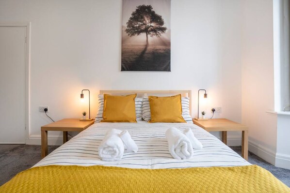A neatly made bed with striped bedding, two pillows, and a yellow throw blanket. There are two bedside tables with lamps, a wall-mounted painting of a tree, and two towels on the bed. - Quiet Neighborhood-free Parking (Blackpool)