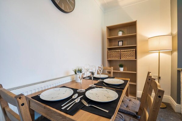 A dining area with a wooden table set for four, equipped with plates, cutlery, and wine glasses. A bookshelf with decorative baskets is against the wall, and a floor lamp is visible to the right. - Quiet Neighborhood-free Parking (Blackpool)