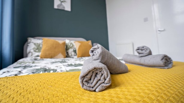 A neatly made bed with a yellow bedspread and rolled towels, a dark blue wall with a small plant, and a white door in a hotel room. - 2 Bed-town Centre-free Parking (Wallsend)