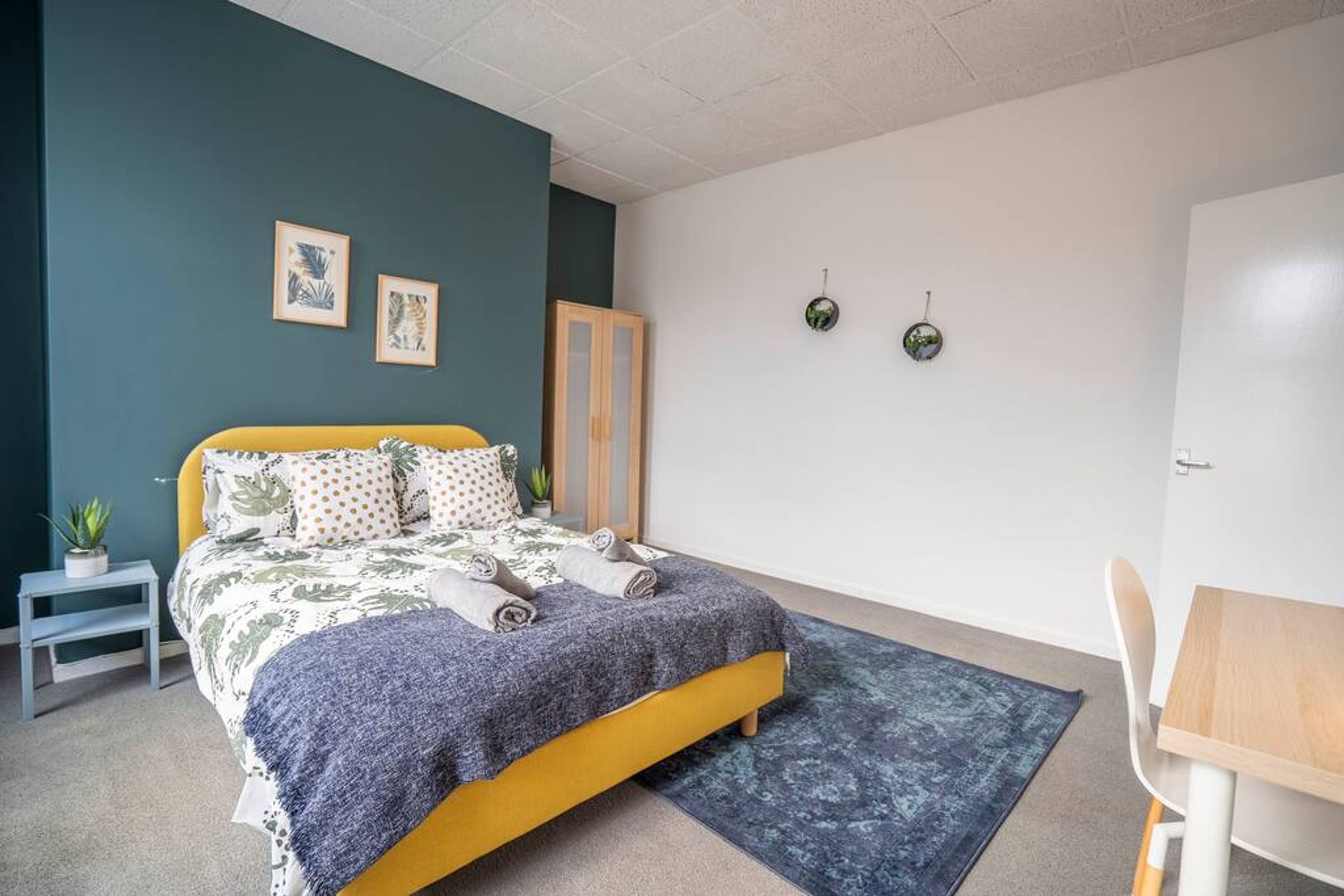 A bedroom with a bed, a small table, a chair, a plant, and two wall-mounted clocks.