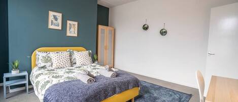 A bedroom with a bed, a small table, a chair, a plant, and two wall-mounted clocks.