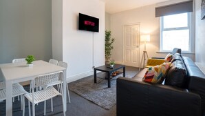 Una sala de estar moderna con una mesa blanca, sillas, un sofá negro con cojines de colores, un televisor montado en la pared y una ventana con vistas.