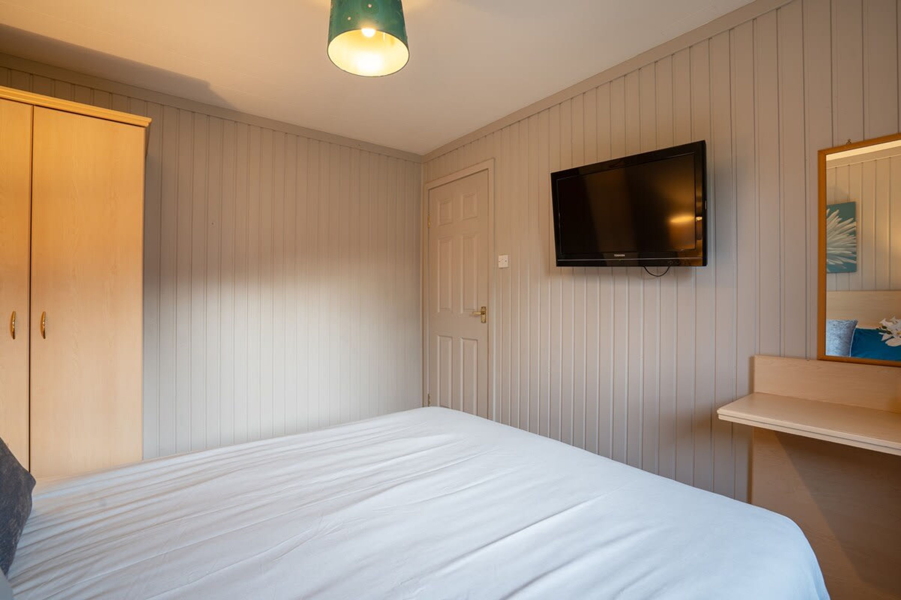 A bedroom with a bed, a wardrobe, a television mounted on the wall, and a mirror above a shelf.