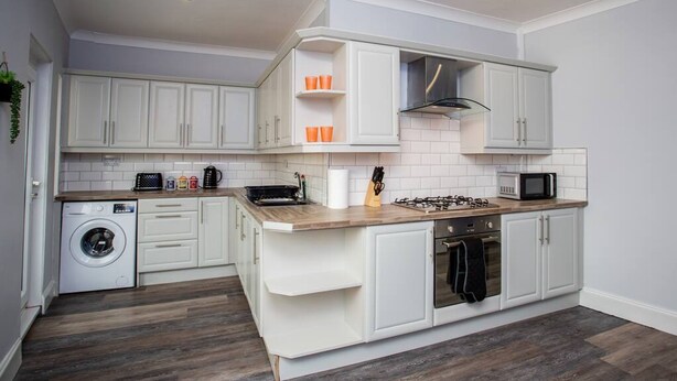 A compact kitchen with white cabinetry, a built-in washing machine, a microwave, and a gas stove. - Large 3 Bed Apartment-sea Road (Sunderland)