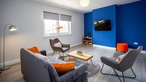 A modern living room with a grey sofa, two armchairs, a coffee table, a floor lamp, a TV on the wall, and a window with blinds. - Large 3 Bed Apartment-sea Road (Sunderland)