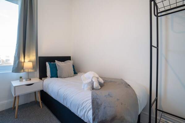 A neatly made bed with a white comforter and a gray blanket, a small bedside table with a lamp, and a window with a curtain. - Cozy 3 Bed Apartment - Parking (Gateshead)