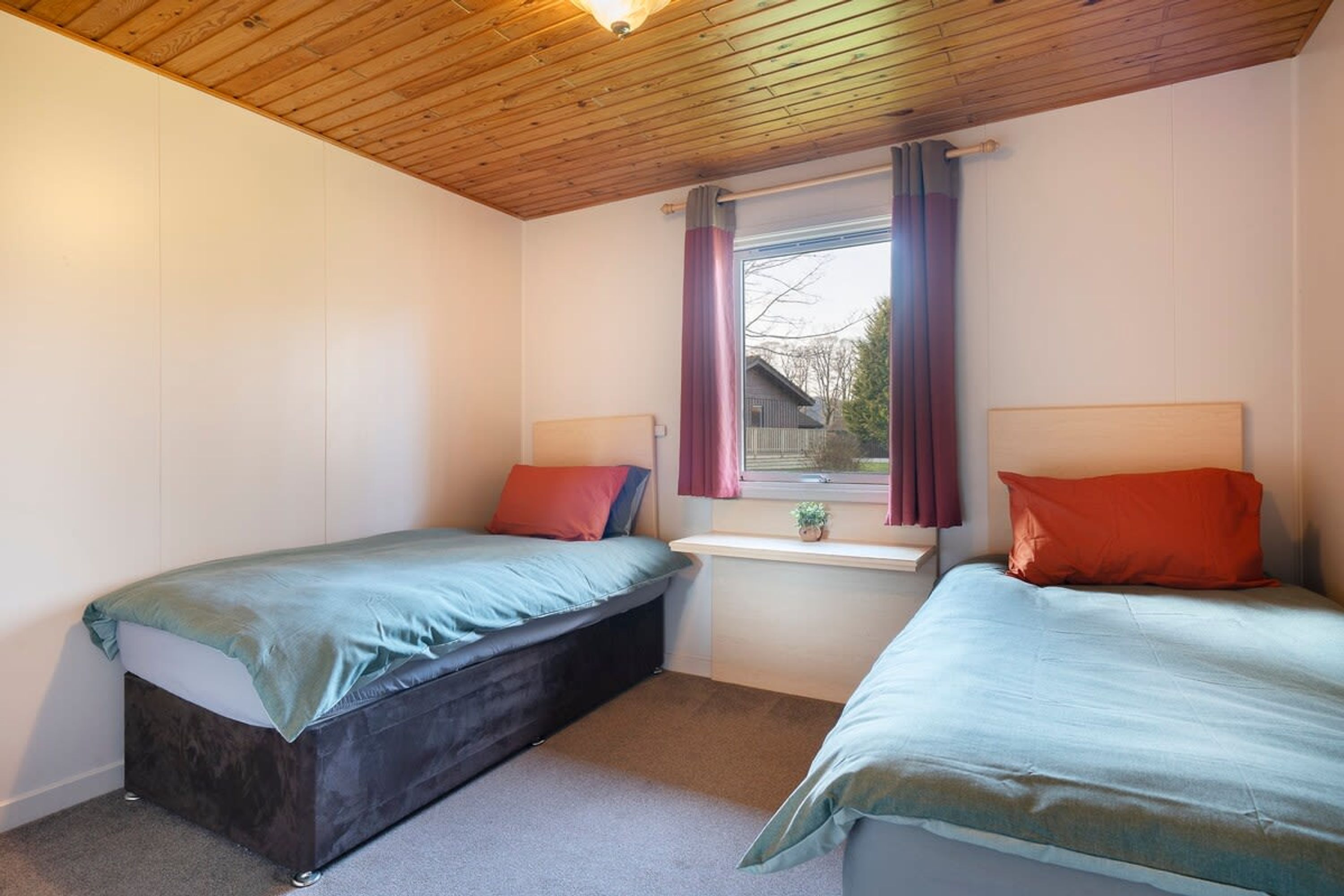 A bedroom with two beds, a window with curtains, a wooden ceiling, and a small table.