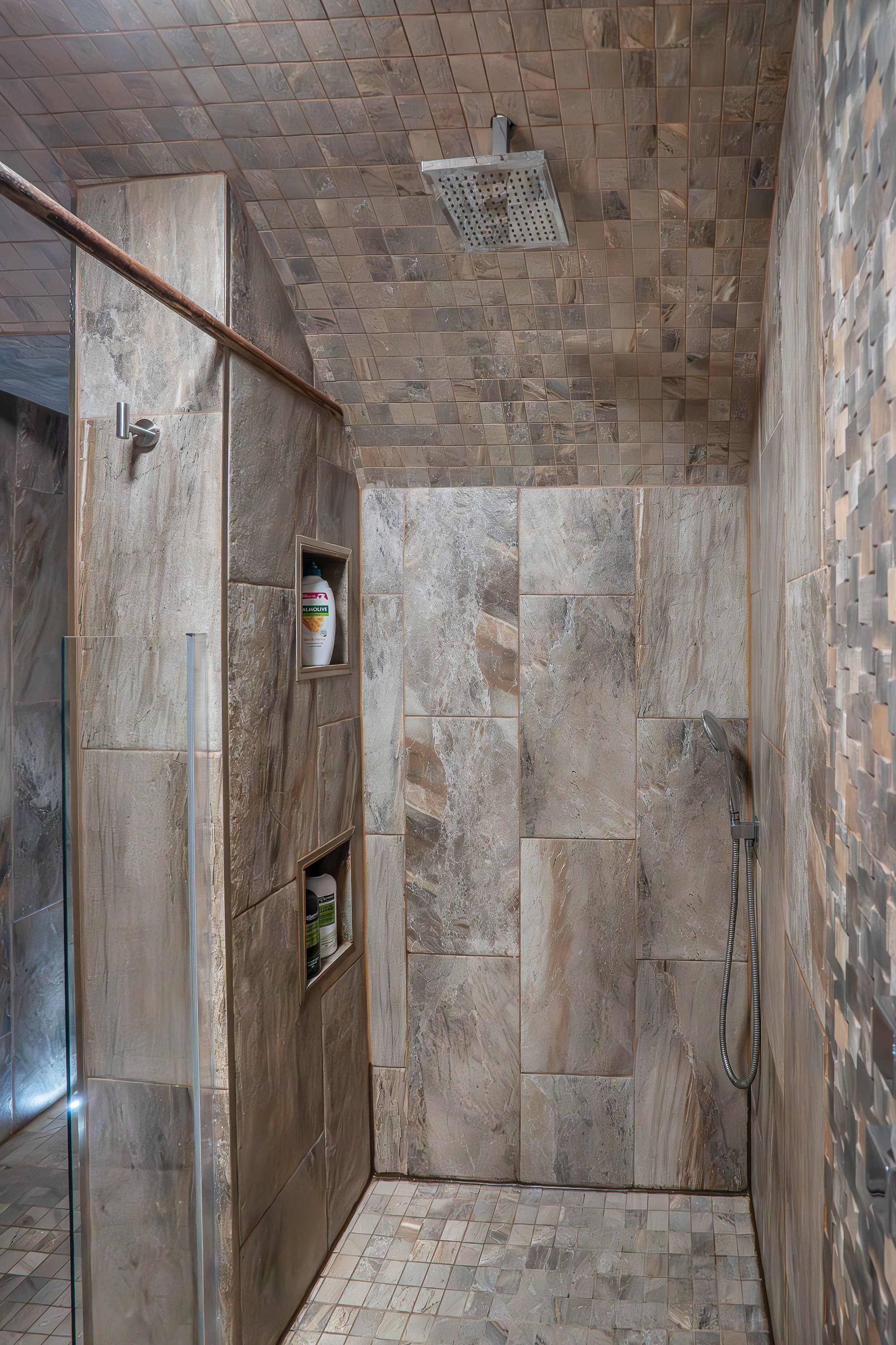 A shower area with a tiled floor and walls, a showerhead, and a shelf with a bottle.