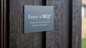 Un letrero en una valla de madera con el texto '¿Se te antoja una parrillada? Por favor, usa el área de barbacoa designada y disfruta.'