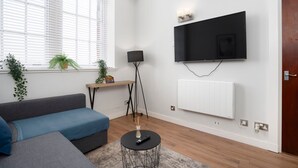 Un salon moderne avec un canapé gris, une petite table ronde noire, un lampadaire et un téléviseur à écran plat fixé au mur.