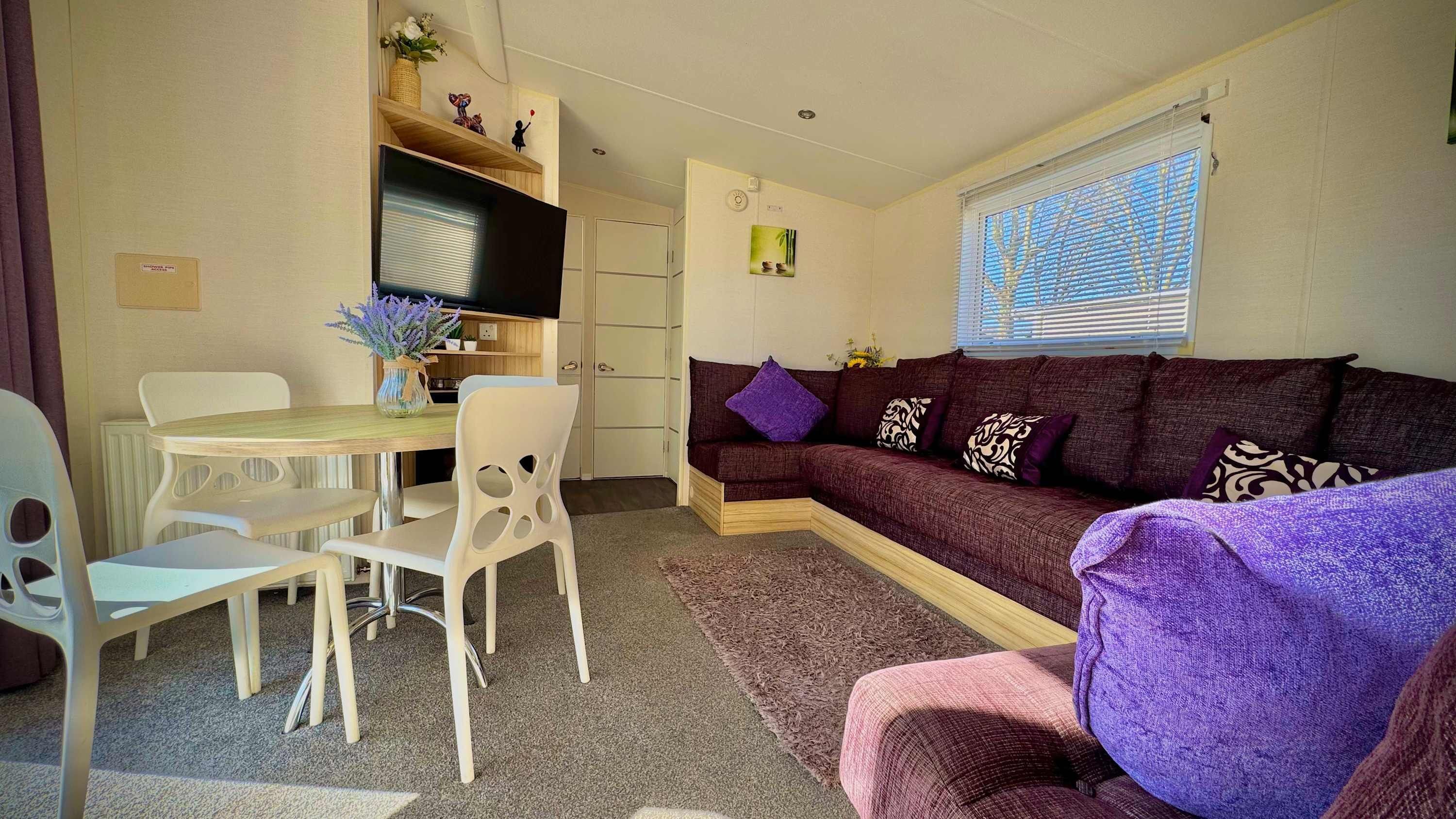 A compact living space with a dining area, a sofa with patterned cushions, and a window with blinds.