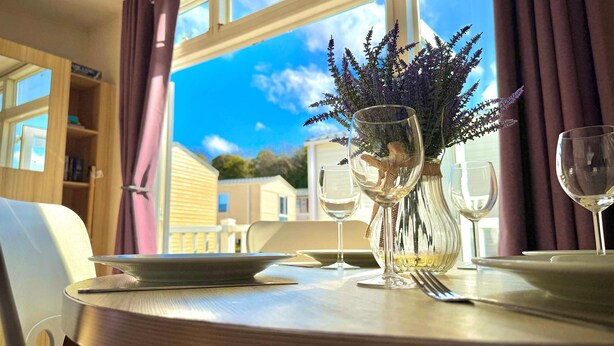 Una mesa de comedor con un florero, dos copas de vino y un plato, frente a una ventana con cortinas moradas.