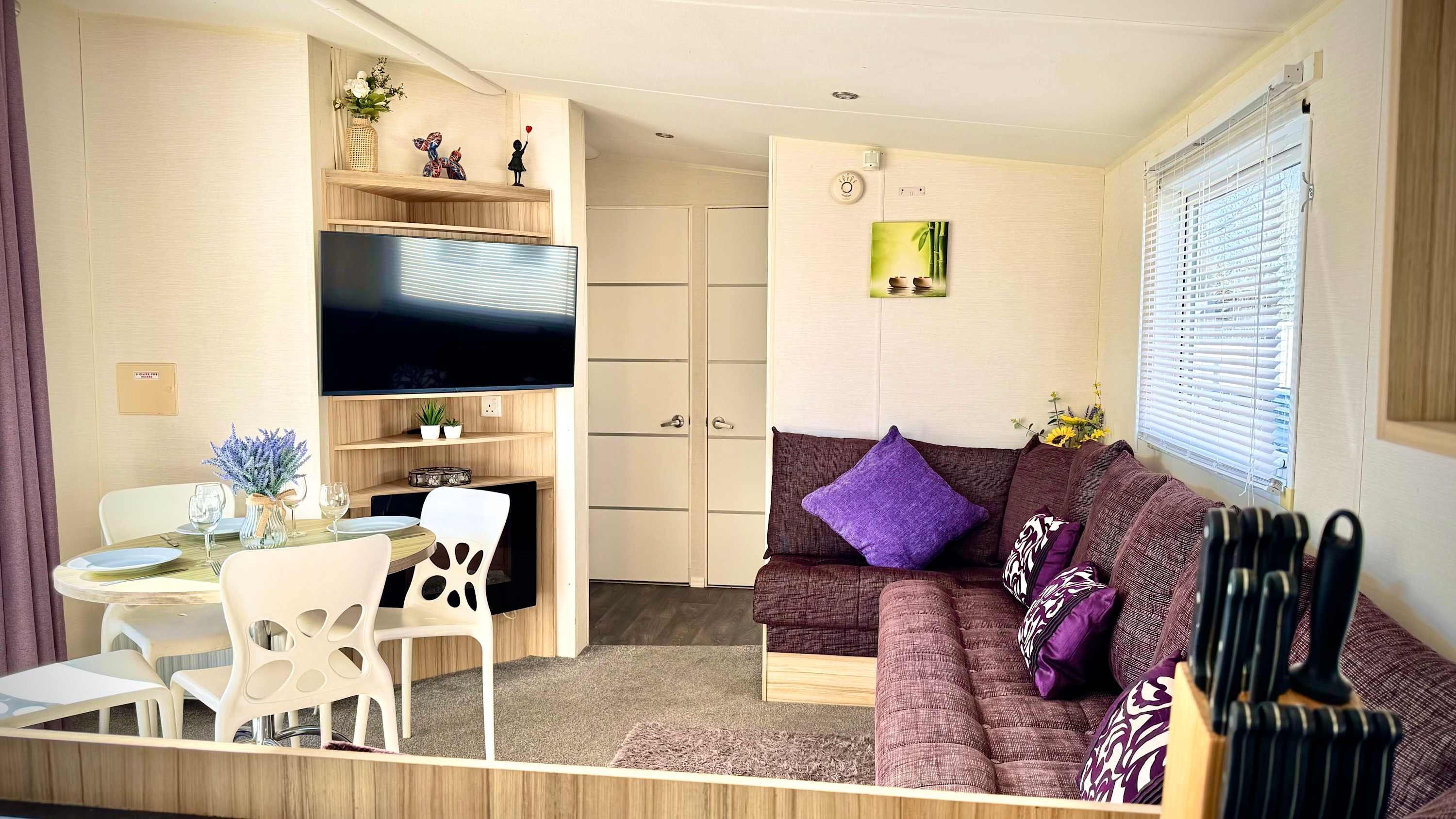 A compact living space with a sofa, a dining table with chairs, a flat-screen TV mounted on the wall, and a small shelf with decorative items.