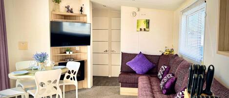A compact living space with a sofa, a dining table with chairs, a flat-screen TV mounted on the wall, and a small shelf with decorative items.
