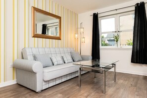 A modern living room with a striped wall, a sofa, a glass coffee table, a mirror, and a window with curtains.