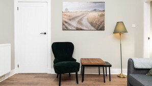 Een kamer met een witte deur, een groene fauteuil, een houten tafel, een vloerlamp en een grijze bank.