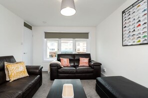 A living room with a black leather sofa, a coffee table, and a large poster of sneakers on the wall. - 3 Bed House-free Parking-6beds (Glasgow)