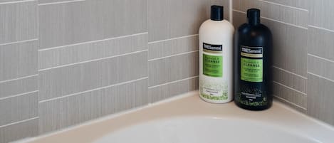 Ein Badezimmer mit Fenster, grauen Fliesenwänden und zwei Flaschen TRESemmé Fruit & Cleanse Shampoo am Badewannenrand.