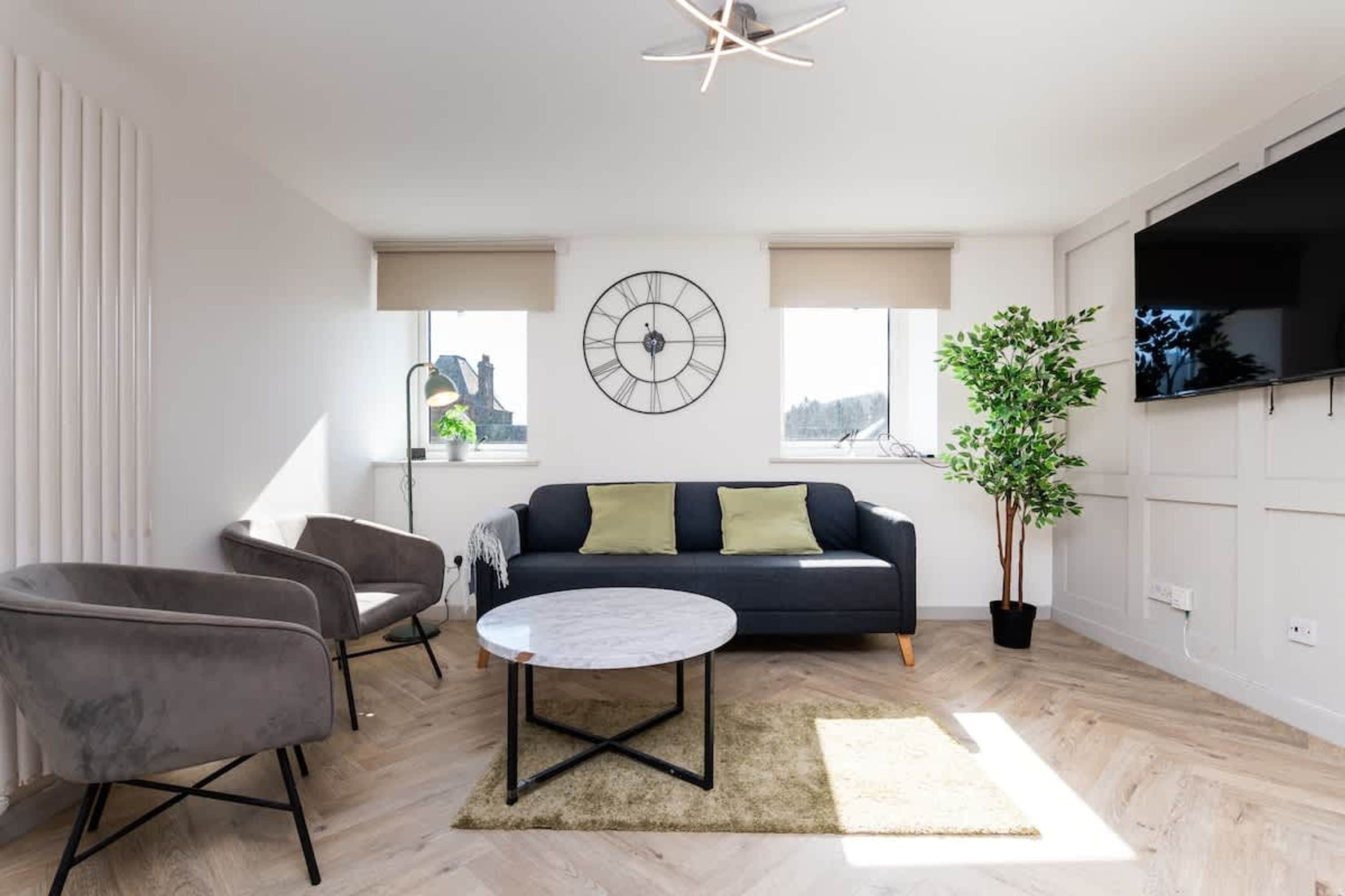 A modern living room with a sofa, two armchairs, a round coffee table, a large wall clock, and a TV mounted on the wall.