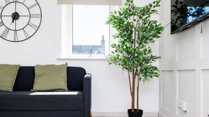 A modern living room with a dark grey sofa, green cushions, a large wall clock, a potted plant, and a window with a view of a cityscape. - 5 Bedroom Apartment - Galashiels (Galashiels)
