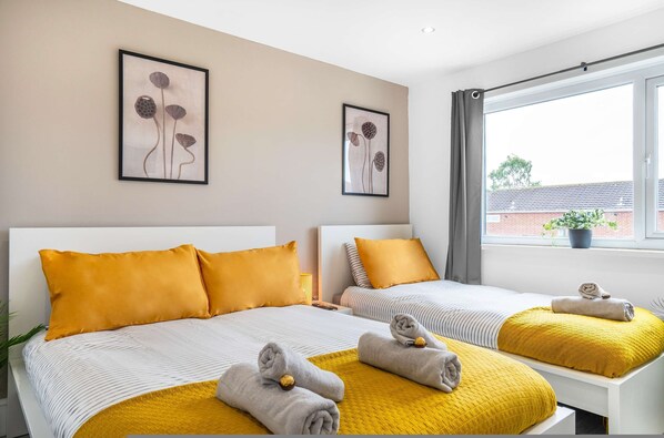 Habitación de hotel con dos camas, cada una con cabecera blanca, ropa de cama amarilla y una toalla enrollada sobre la cama. Hay cuadros con motivos florales en la pared sobre las camas.