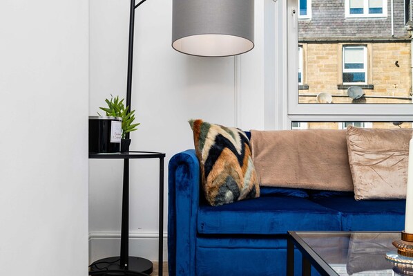 Een moderne woonkamer met een blauwe bank, een zwarte vloerlamp en een glazen salontafel.