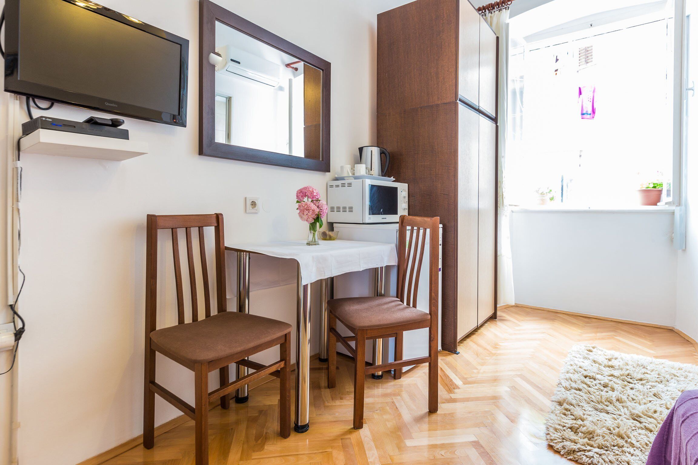 A small dining area with two wooden chairs, a white table, a TV mounted above, and a microwave on the table.