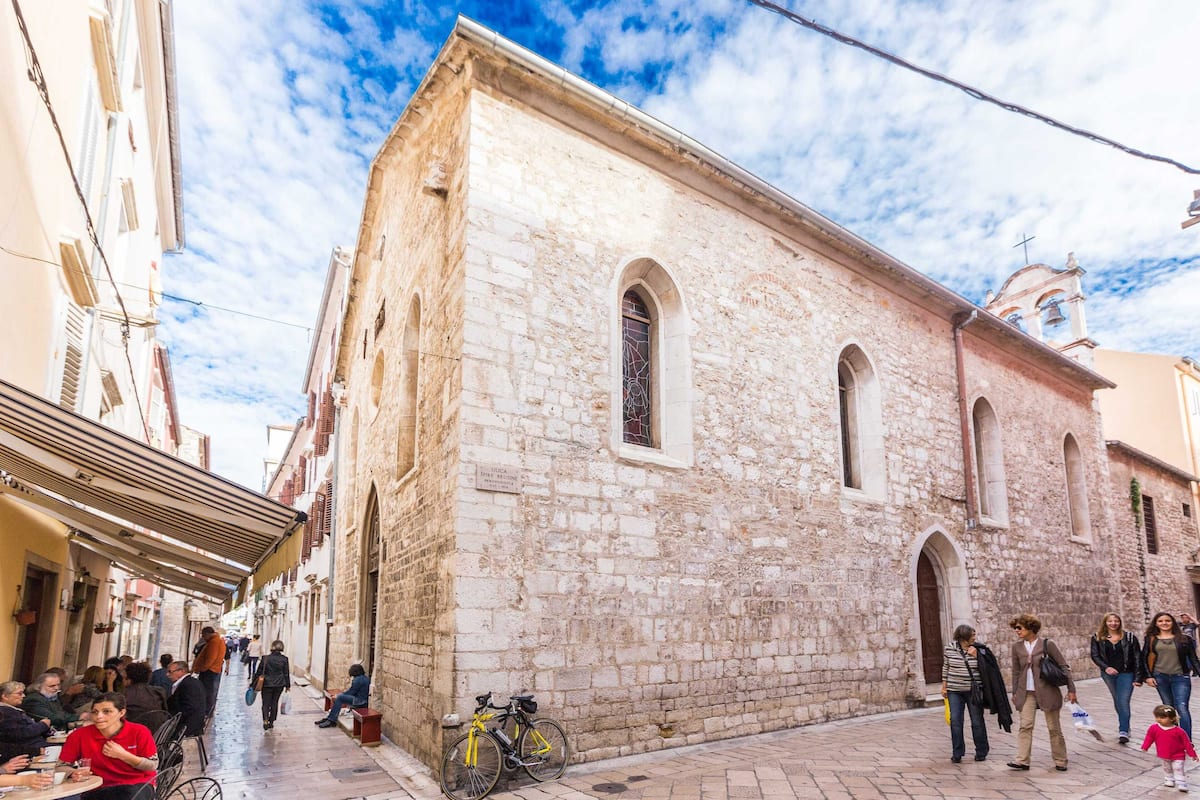 A stone building with arched windows and a bell tower, surrounded by pedestrians and a bicycle.