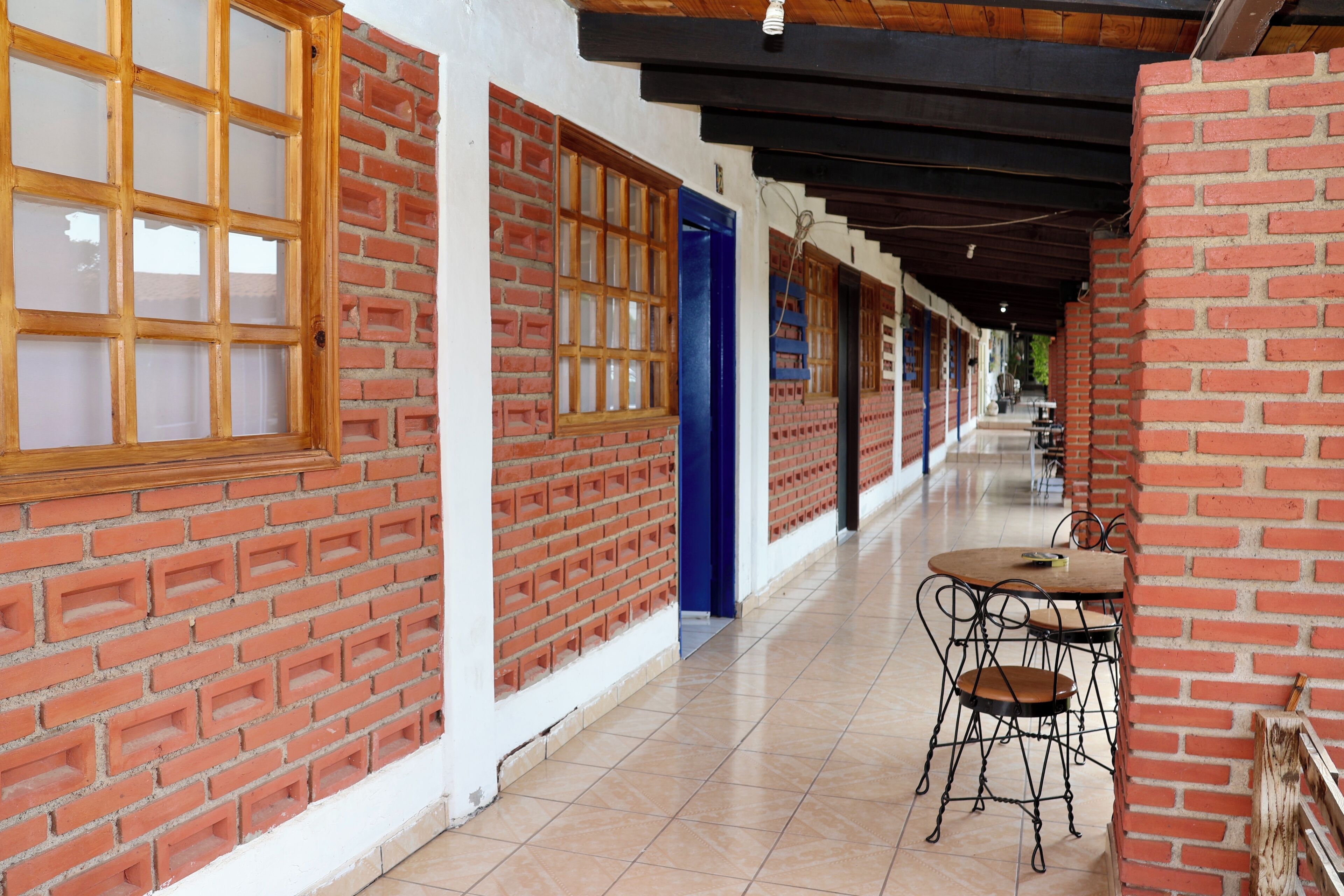A corridor with brick walls, wooden-framed windows, and a blue door. There are metal chairs and a small round table in the corridor.
