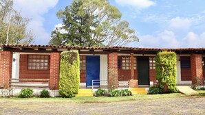 A two-story brick building with a tiled roof, a blue door, and a small front porch with a bench. - La Palma, Suites Campestres (Jocotitlán)