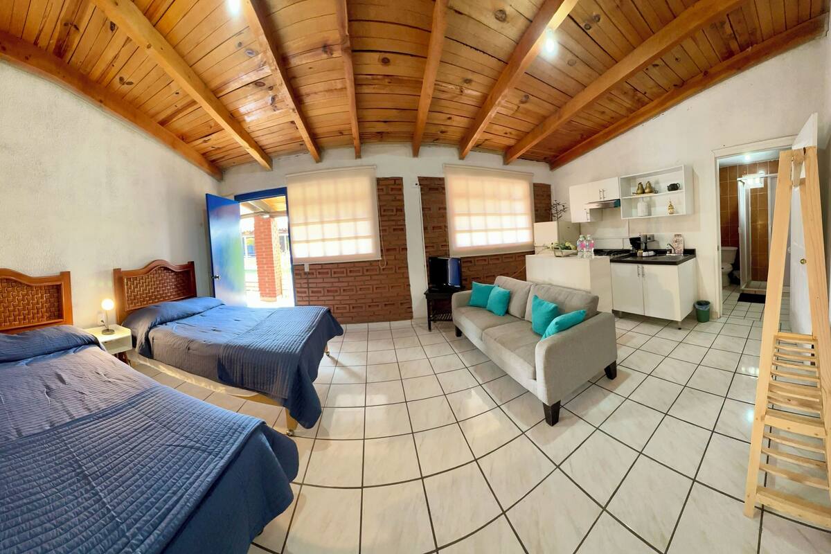 A room with two beds, a sofa, a wooden ladder, and a kitchen area.