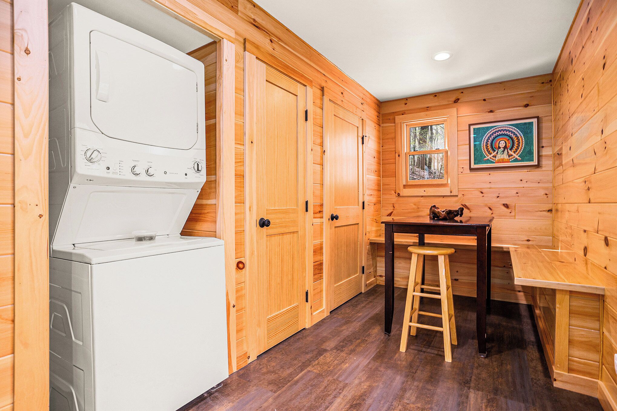 A compact kitchen with wooden walls, a washing machine, a wooden table with a stool, and a window with a view of the forest.